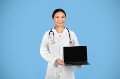 Smiling Asian Doctor Woman In White Coat Holding Laptop With Blank Screen, Beautiful Female Therapist Recommending Website For Online Medical Consultancy, Standing On Blue Background, Mockup